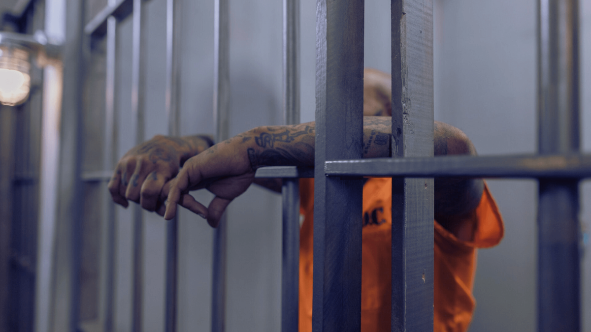 Inmate in orange jumpsuit gripping prison cell bars with tattooed hands.