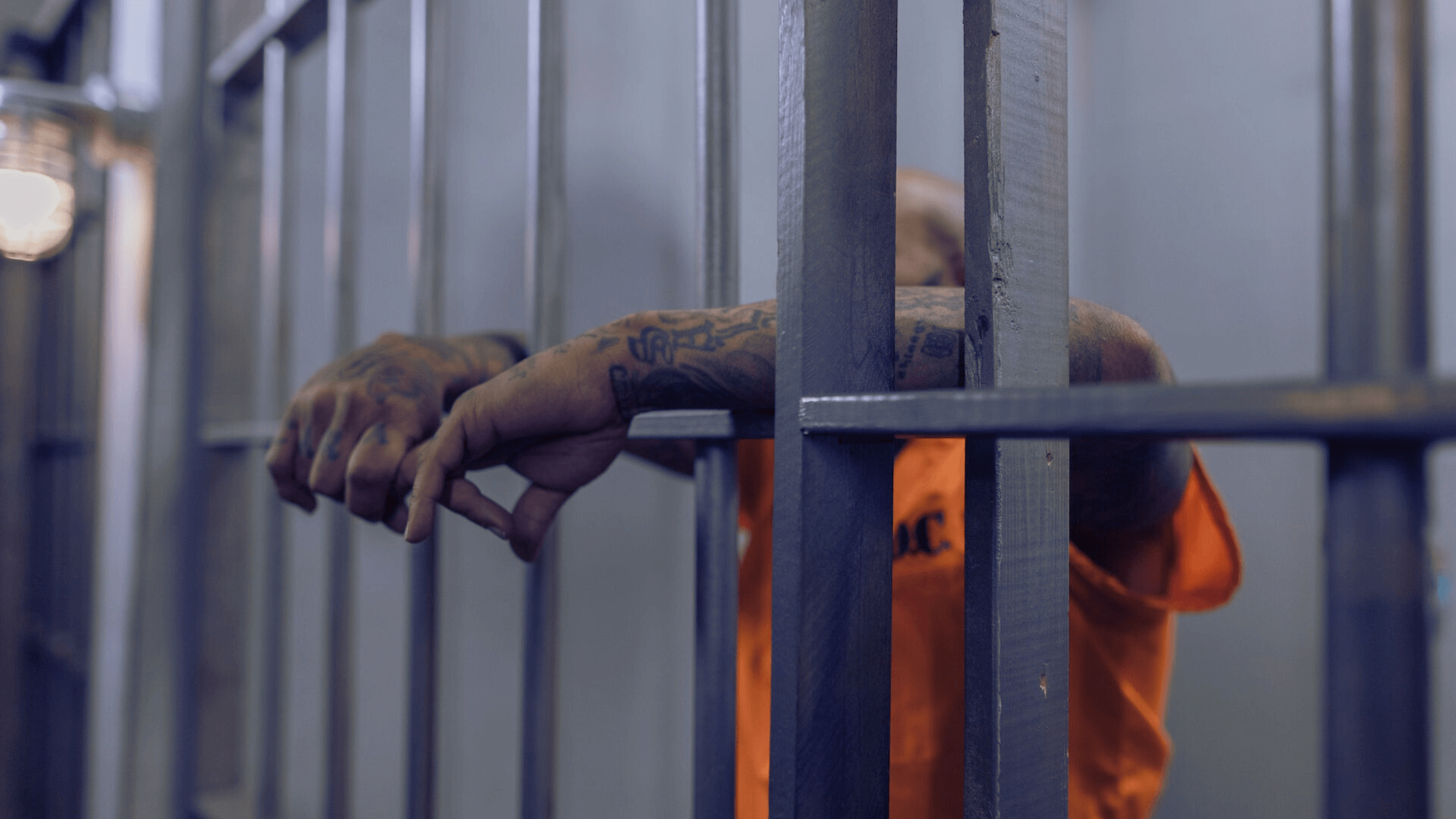 Inmate in orange jumpsuit gripping prison cell bars.